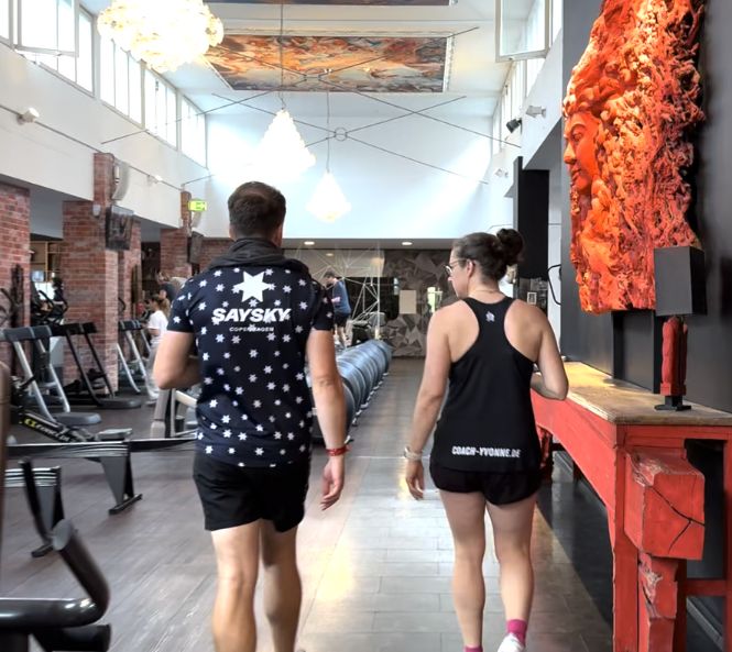 Zwei Personen, eine mit einem "SAYSKY"-Shirt und die andere mit einem "COACH-YVONNE"-Shirt, gehen in einem Fitnessstudio nebeneinander her.