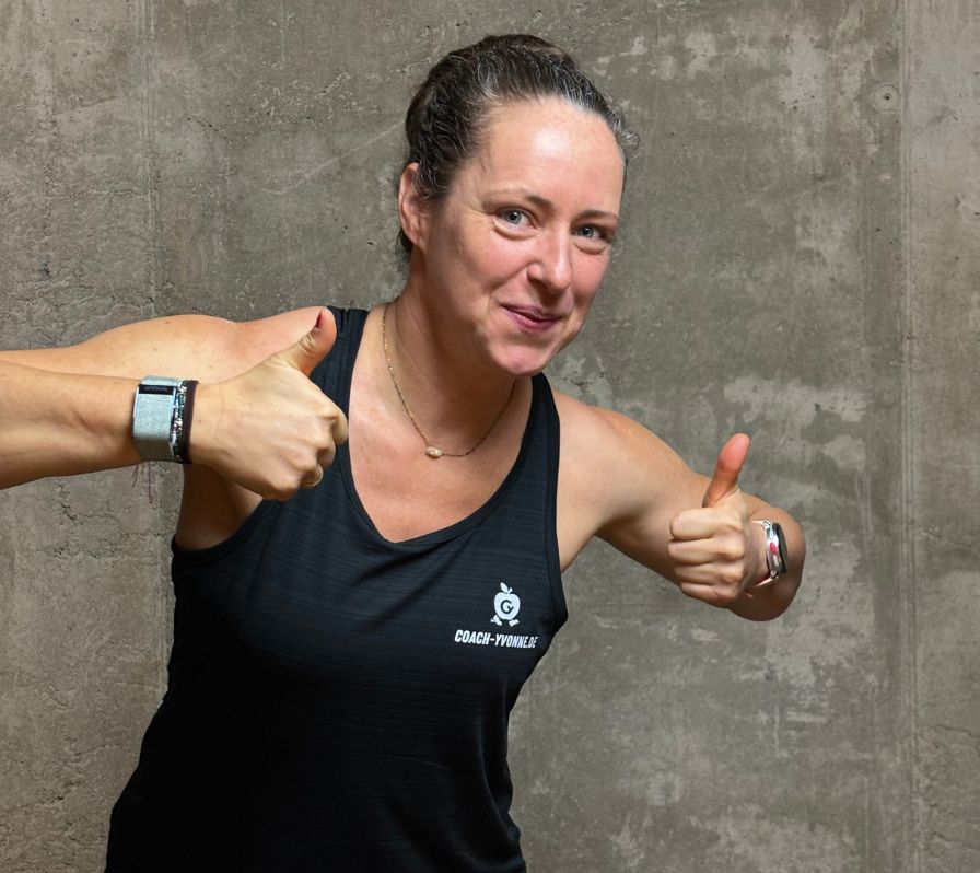 Eine Frau in einem schwarzen Sportshirt mit dem Text "COACH-YVONNE.DE" zeigt mit beiden Daumen nach oben.