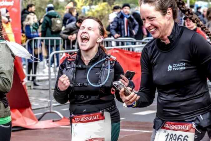 Zwei Frauen feiern am Ziel einer Laufveranstaltung, eine jubelt laut, die andere hält ein Smartphone und lächelt.