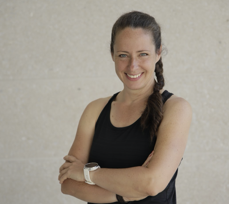 Eine Frau mit geflochtenem Haar und schwarzer Sportbekleidung steht lächelnd mit verschränkten Armen vor einem hellen Hintergrund.